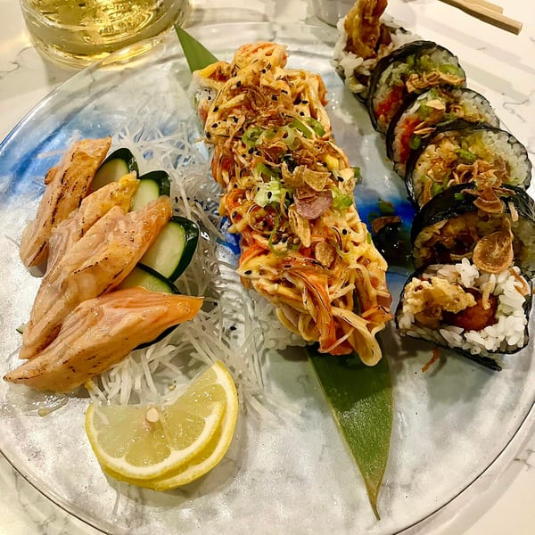 A plate of sushi rolls, seared salmon sashimi, and a lemon wedge, with a drink in the background.