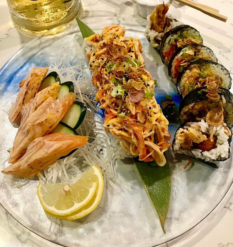 A plate of sushi rolls, seared salmon sashimi, and a lemon wedge, with a drink in the background.