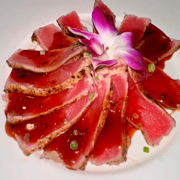 Seared tuna sashimi slices with red sauce and a purple orchid garnish on a white plate.