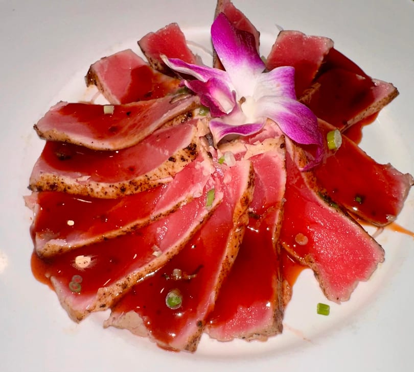 Seared tuna sashimi slices with red sauce and a purple orchid garnish on a white plate.