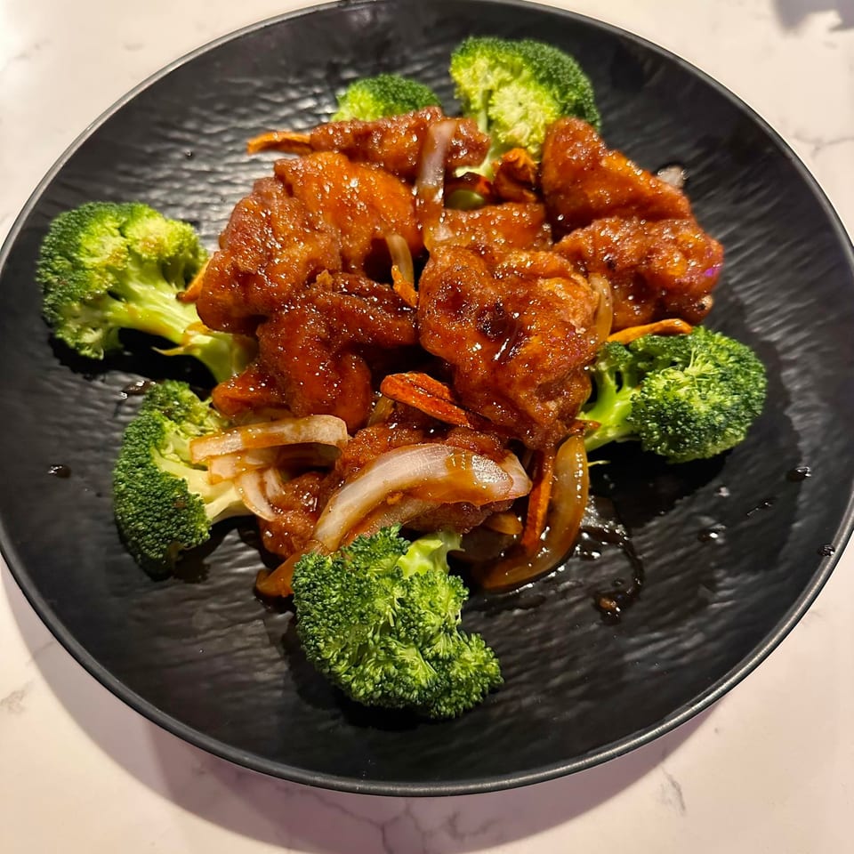 Sweet and sour chicken with broccoli and onions on a black plate.