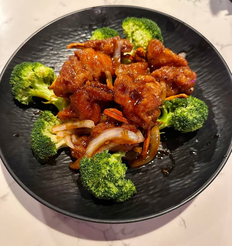 Sweet and sour chicken with broccoli and onions on a black plate.